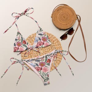 May include: A floral print bikini with a triangle top and a high-cut bottom. The bikini is on a woven straw mat with a round straw bag and sunglasses.