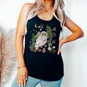 May include: Black vest top with a detailed illustration of a white owl surrounded by botanical elements. The design includes ferns, flowers, a crescent moon, and a dragonfly. The owl has yellow eyes and is perched among the foliage.