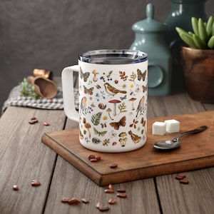 Può includere: Tazza in ceramica bianca con un motivo colorato di uccelli, farfalle, funghi e fiori. La tazza ha un manico nero e un coperchio blu.