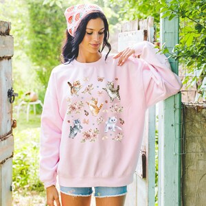 May include: A pink sweatshirt with a graphic print of playful kittens and flowers. The kittens are in various poses, some are jumping, some are sitting, and some are standing. The flowers are in shades of pink, white, and yellow.