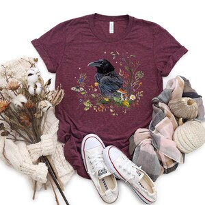 May include: A maroon t-shirt with a graphic of a black raven surrounded by flowers and mushrooms. The raven is perched on a branch with its wings spread. The shirt is made of a soft, comfortable material and is perfect for everyday wear.