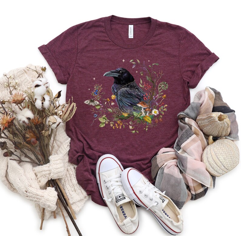 May include: A maroon t-shirt with a graphic of a black raven surrounded by flowers and mushrooms. The raven is perched on a branch with its wings spread. The shirt is made of a soft, comfortable material and is perfect for everyday wear.