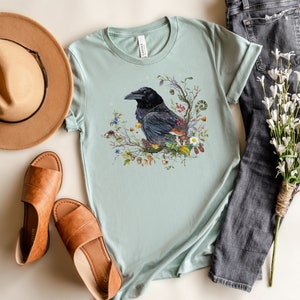 May include: A light blue t-shirt with a black crow surrounded by flowers and leaves. The crow is perched on a branch with a mushroom and other forest elements.