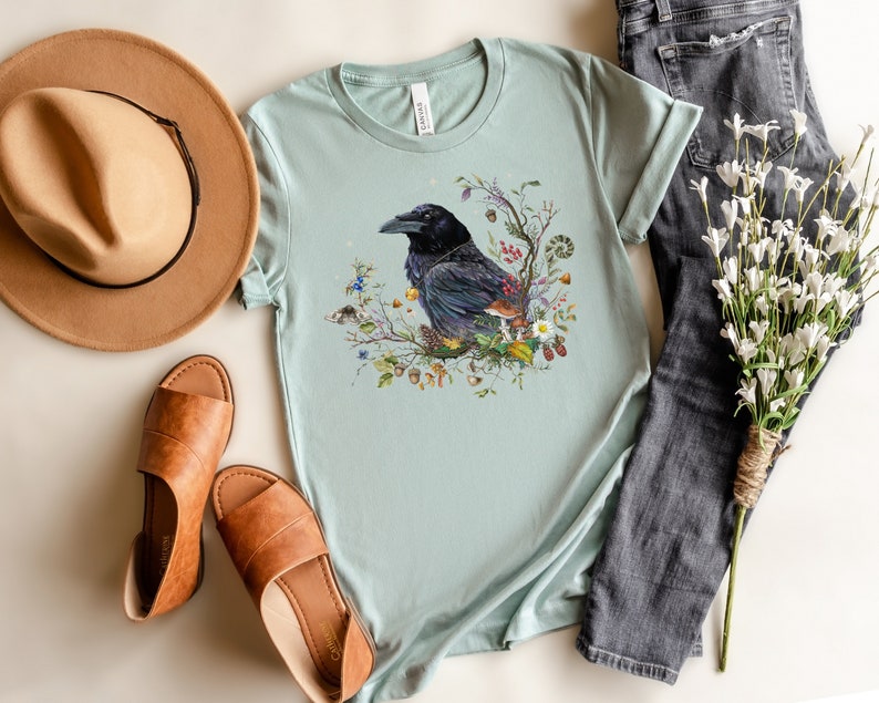 May include: A light blue t-shirt with a black crow surrounded by flowers and leaves. The crow is perched on a branch with a mushroom and other forest elements.