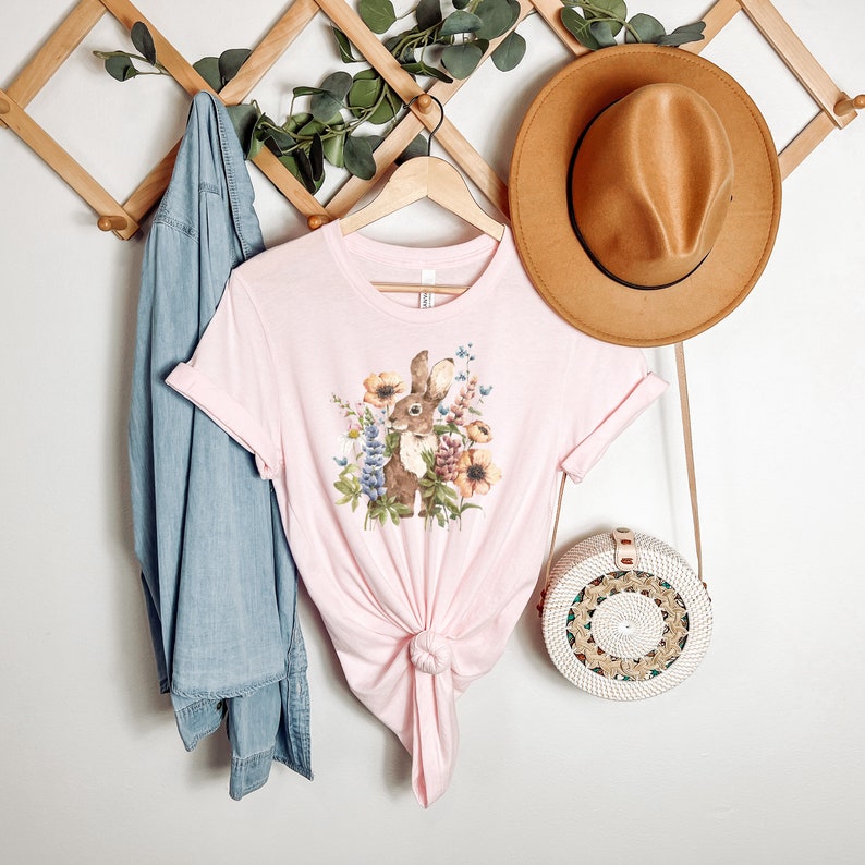 Op de afbeelding: Een roze T-shirt met een afbeelding van een bruin konijn omringd door bloemen. Het konijn kijkt naar links. Het shirt is onderaan geknoopt.