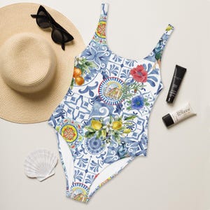 May include: One-piece swimsuit with a blue and white tile pattern, featuring lemons, oranges, and floral accents. A straw hat, sunglasses, sunscreen, and a seashell are also visible.