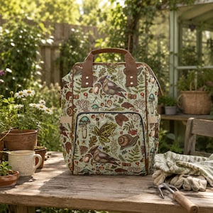 Könnte beinhalten: Ein hellgrüner Rucksack mit einem Naturmuster mit Vögeln, Pilzen und Blättern. Der Rucksack hat braune Ledergriffe und Akzente. Er steht auf einem Holztisch mit Gartengeräten und Pflanzen im Hintergrund.