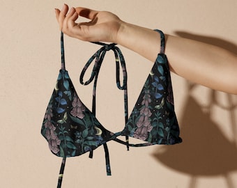 Op de afbeelding: Een zwarte driehoekige bikini top met een bloemenprint met maan- en vlindermotieven. De print is donkergroen, paars en wit op een zwarte achtergrond.