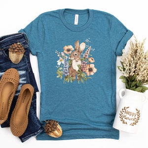 Op de afbeelding: Een tealblauw T-shirt met een aquarel-illustratie van een bruin konijn omringd door bloemen. De bloemen zijn roze, paars, wit en geel. Het shirt ligt plat op een witte achtergrond.