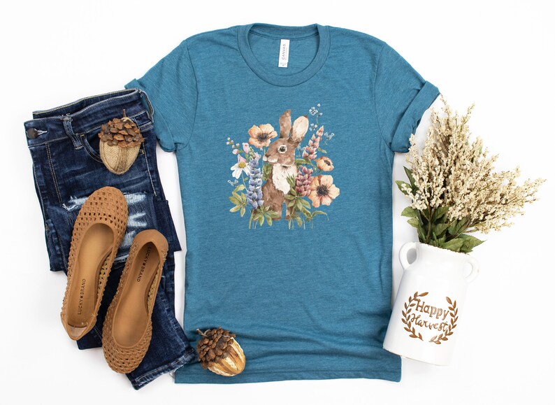 Op de afbeelding: Een tealblauw T-shirt met een aquarel-illustratie van een bruin konijn omringd door bloemen. De bloemen zijn roze, paars, wit en geel. Het shirt ligt plat op een witte achtergrond.