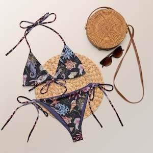 Könnte beinhalten: Ein blauer und lilafarbener Bikini mit einem Muschel- und Seepferdchen-Druck. Das Bikinioberteil ist ein Dreieck-Stil mit verstellbaren Trägern. Das Bikiniunterteil ist ein Binde-Seitenstil. Der Bikini liegt auf einer geflochtenen Strohhütte.