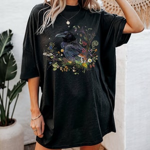 May include: Black oversized t-shirt with a graphic print of a raven surrounded by flowers, mushrooms, and leaves.