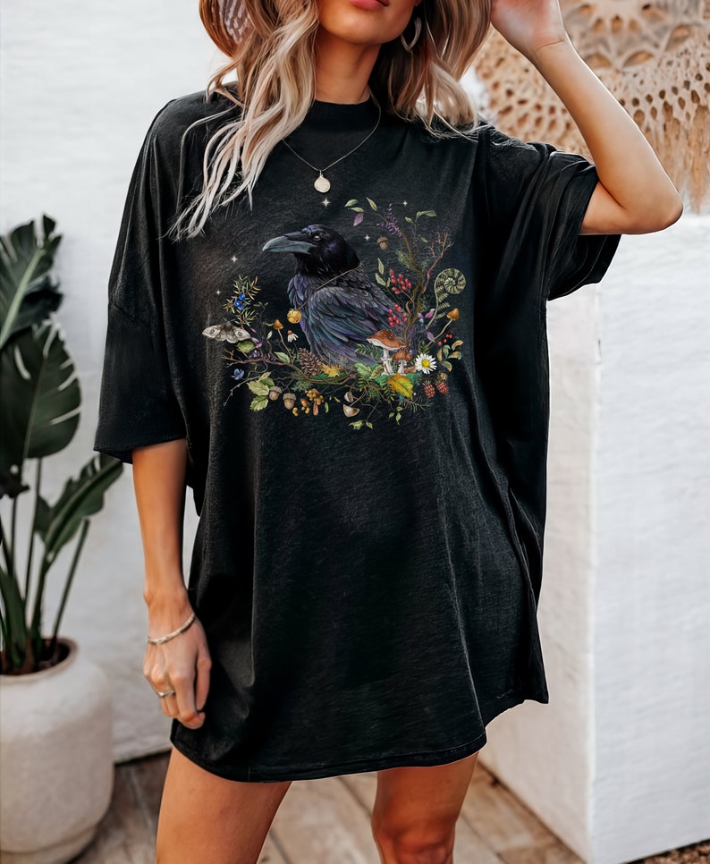 May include: Black oversized t-shirt with a graphic print of a raven surrounded by flowers, mushrooms, and leaves.