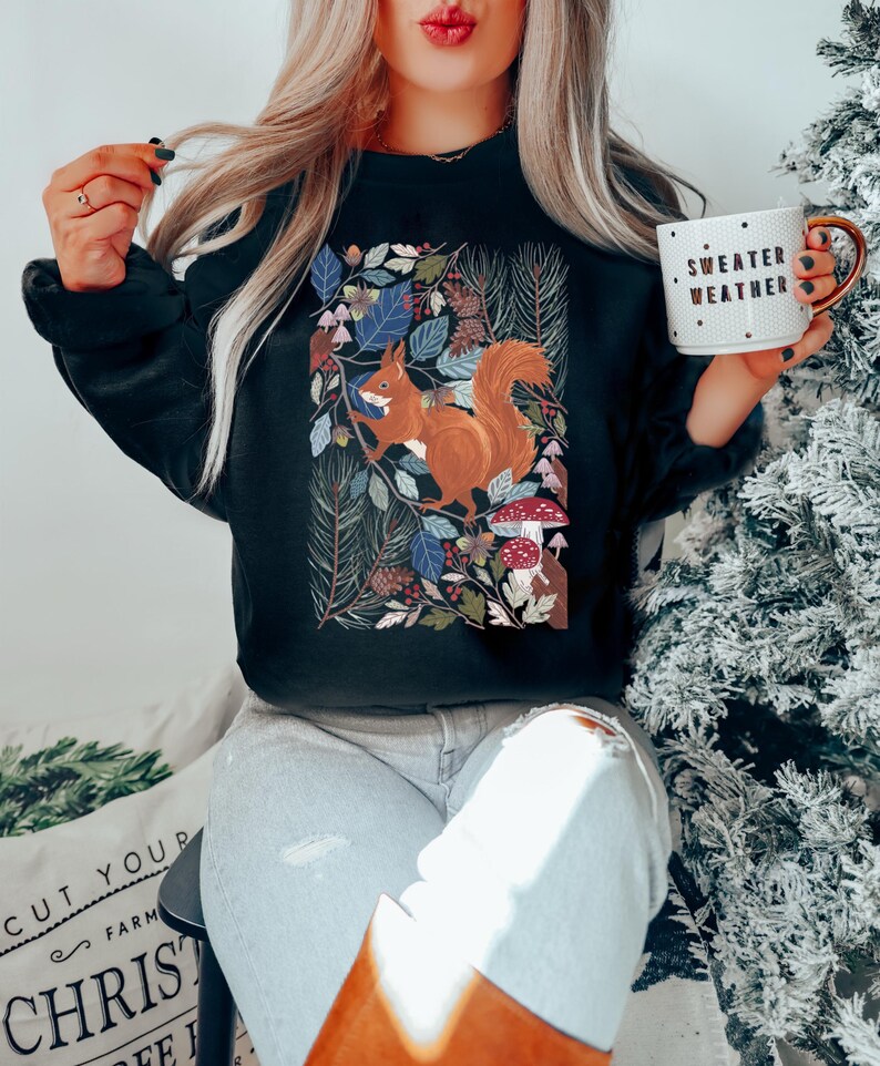 May include: Black sweatshirt featuring a colourful illustration of a squirrel in a woodland scene. The text "Sweater Weather" is printed on a white mug in the image.