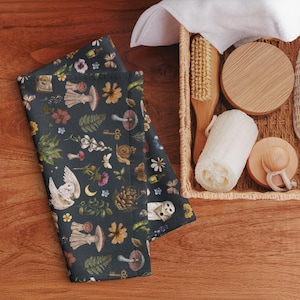 Op de afbeelding: Donkerblauwe gevouwen handdoek met een grillig bospatroon met paddenstoelen, uilen, sleutels en bloemen. De handdoek ligt naast een rieten mand met een houten borstel, een luffa en houten deksels, alles op een houten oppervlak.
