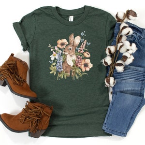 Op de afbeelding: Een groen T-shirt met een aquarel-illustratie van een bruin konijn omringd door kleurrijke bloemen. Het shirt wordt gecombineerd met bruine su&egrave;de laarzen met franjes en een blauwe jeans.