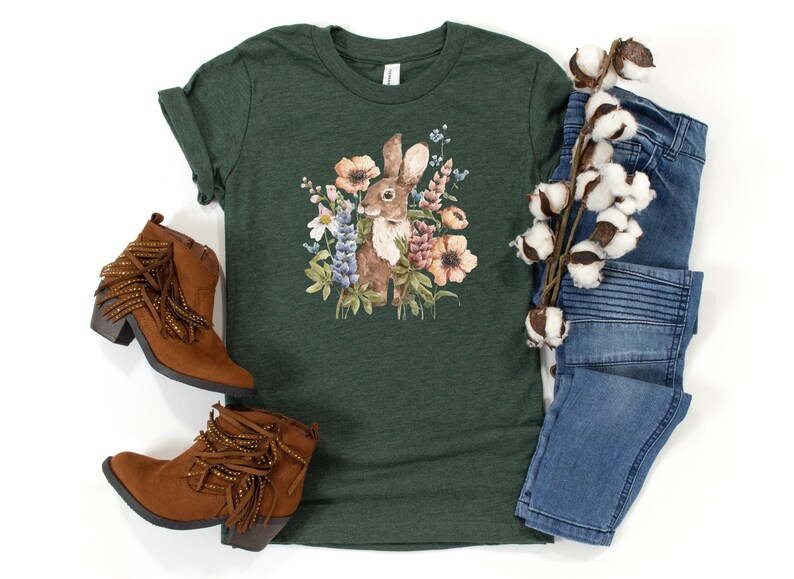 Op de afbeelding: Een groen T-shirt met een aquarel-illustratie van een bruin konijn omringd door kleurrijke bloemen. Het shirt wordt gecombineerd met bruine su&egrave;de laarzen met franjes en een blauwe jeans.