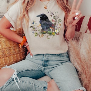 May include: A beige t-shirt with a graphic of a black raven surrounded by flowers, leaves, and berries.