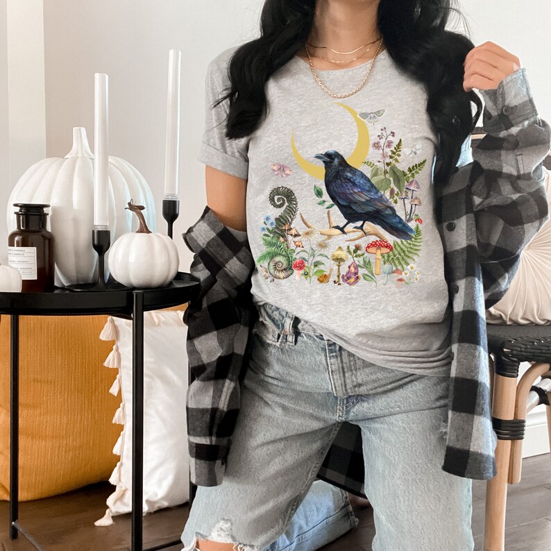 May include: A gray t-shirt with a graphic of a black crow perched on a crescent moon surrounded by flowers and mushrooms.