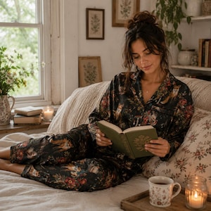 Peut inclure: Une personne portant un ensemble de pyjama noir avec un imprimé d'animaux de la forêt et de fleurs, lisant un livre sur un lit. La pièce a une esthétique chaleureuse et naturelle avec une fenêtre, des bougies et une tasse à fleurs.