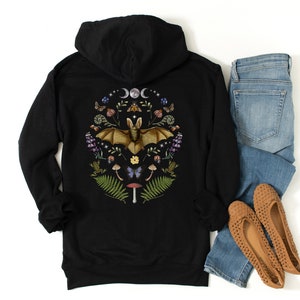 May include: Black hooded sweatshirt featuring a graphic print of a bat surrounded by flowers, mushrooms, and other woodland elements. The graphic is in shades of brown, green, purple, and yellow.