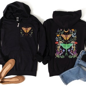 May include: Two black zip-up hoodies with a moth and floral design. The hoodie on the left has a single moth on the front, while the hoodie on the right has a larger design with two moths and flowers on the back.