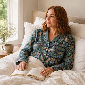 May include: A woman wearing a dark blue pajama top with a floral and bird pattern, and green trim, is sitting in bed with a book. The pajamas are button-down and long-sleeved. The bed has white linens and pillows.