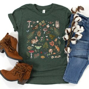 May include: A dark green t-shirt with a colorful illustration of woodland creatures, mushrooms, and plants. The design features a rabbit, owl, mouse, butterflies, and more.