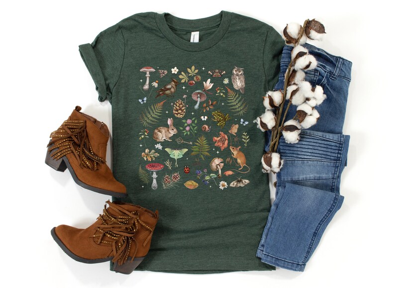 May include: A dark green t-shirt with a colorful illustration of woodland creatures, mushrooms, and plants. The design features a rabbit, owl, mouse, butterflies, and more.
