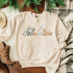 May include: A beige sweatshirt featuring a watercolour design of woodland animals, including a raccoon, a deer, a rabbit, and a squirrel. The animals are surrounded by flowers and leaves.