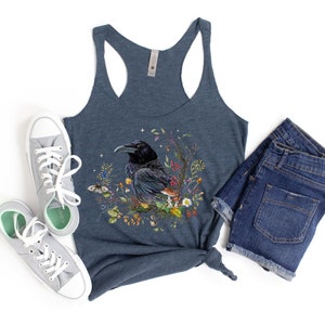K&ouml;nnte beinhalten: Ein dunkelblaues Tanktop mit einem Aufdruck eines schwarzen Rabens, umgeben von Blumen und Pilzen. Das Tanktop ist aus einem weichen, bequemen Material gefertigt.