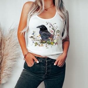K&ouml;nnte beinhalten: Ein wei&szlig;es Tanktop mit einer schwarzen Kr&auml;he Illustration, umgeben von Blumen und Bl&auml;ttern. Die Kr&auml;he sitzt auf einem Ast mit Beeren und Eicheln.