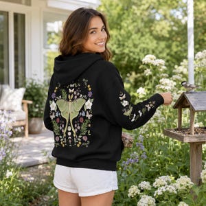 May include: A black hooded sweatshirt featuring a large, detailed graphic on the back of a pale green luna moth surrounded by colorful wildflowers, ferns, and small butterflies. The right sleeve also displays a matching floral and moth print.