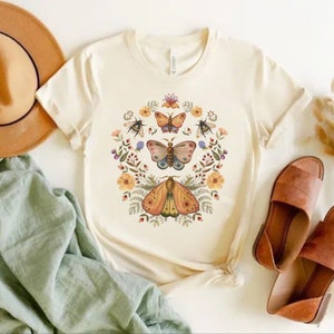 May include: A cream-colored short-sleeve t-shirt featuring a central graphic print of three butterflies, two bees, and various colorful flowers and foliage arranged in a circular wreath pattern on the front of the garment.