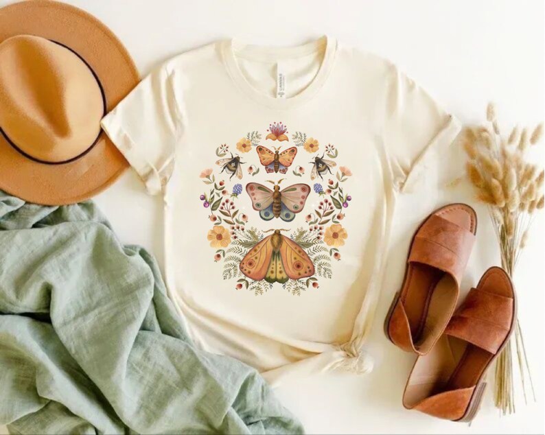 May include: A cream-colored short-sleeve t-shirt featuring a central graphic print of three butterflies, two bees, and various colorful flowers and foliage arranged in a circular wreath pattern on the front of the garment.