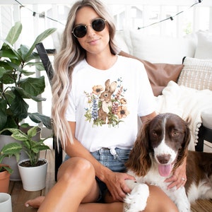 Op de afbeelding: Een wit T-shirt met een aquarel-illustratie van een bruin konijn omringd door bloemen. Het konijn kijkt naar links en heeft lange oren. De bloemen zijn in tinten roze, geel en blauw. Een persoon draagt het T-shirt en zit op een veranda met een bruin-witte hond.