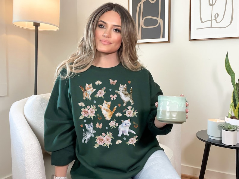 May include: A dark green sweatshirt with a print of kittens and butterflies. The sweatshirt has a crew neck and long sleeves.