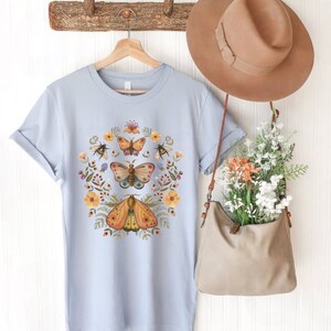 May include: A light blue t-shirt featuring a central graphic of three moths surrounded by a circular arrangement of yellow flowers, green foliage, and small bees. A tan hat and a tan shoulder bag filled with flowers hang nearby.