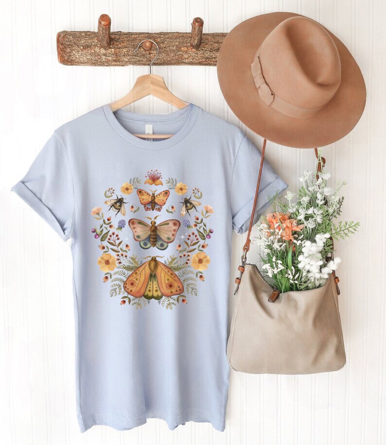 May include: A light blue t-shirt featuring a central graphic of three moths surrounded by a circular arrangement of yellow flowers, green foliage, and small bees. A tan hat and a tan shoulder bag filled with flowers hang nearby.