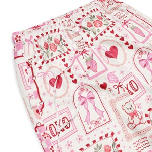 May include: White pajama pants with a repeating pattern of pink hearts, roses, strawberries, bows, and teddy bears. The drawstring waist is visible. The overall design is romantic and feminine.