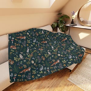 May include: A dark teal throw blanket with a woodland pattern featuring foxes, owls, rabbits, squirrels, mushrooms, and flowers. The blanket is draped over a white sofa, showcasing the detailed nature-themed design.