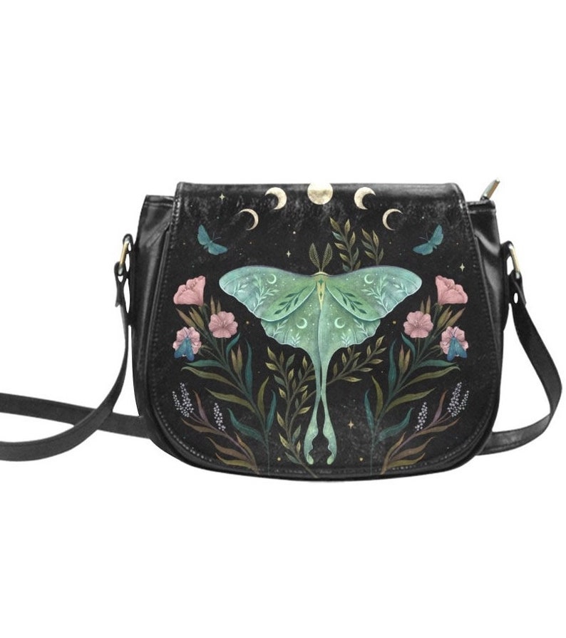 K&ouml;nnte beinhalten: Schwarze Leder-Umh&auml;ngetasche mit Mond- und Mottenmotiv. Die Motte ist gr&uuml;n und hat ein Blumenmuster. Die Tasche hat einen langen verstellbaren Riemen.