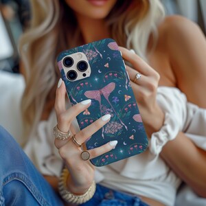May include: A phone case with a dark blue background and a pink moth design. The case has a floral pattern with pink, white, and green accents. The case is designed for an iPhone.