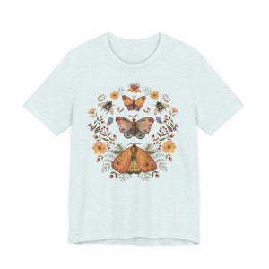 May include: A light heather blue t-shirt featuring a central graphic print of three moths arranged vertically, surrounded by a circular wreath of orange flowers, green leaves, and small berries. The design is symmetrical and detailed in earthy tones.