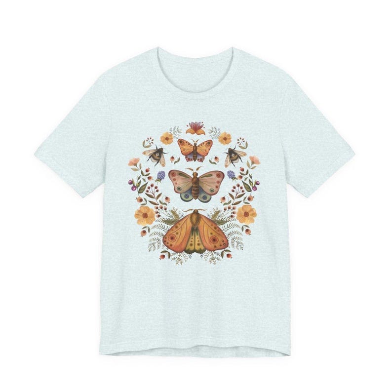 May include: A light heather blue t-shirt featuring a central graphic print of three moths arranged vertically, surrounded by a circular wreath of orange flowers, green leaves, and small berries. The design is symmetrical and detailed in earthy tones.