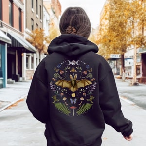 May include: Black hoodie featuring a printed design of a bat surrounded by flowers, mushrooms, and other woodland elements. The graphic also includes moons and stars.