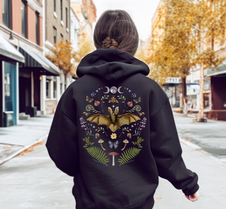 May include: Black hoodie featuring a printed design of a bat surrounded by flowers, mushrooms, and other woodland elements. The graphic also includes moons and stars.