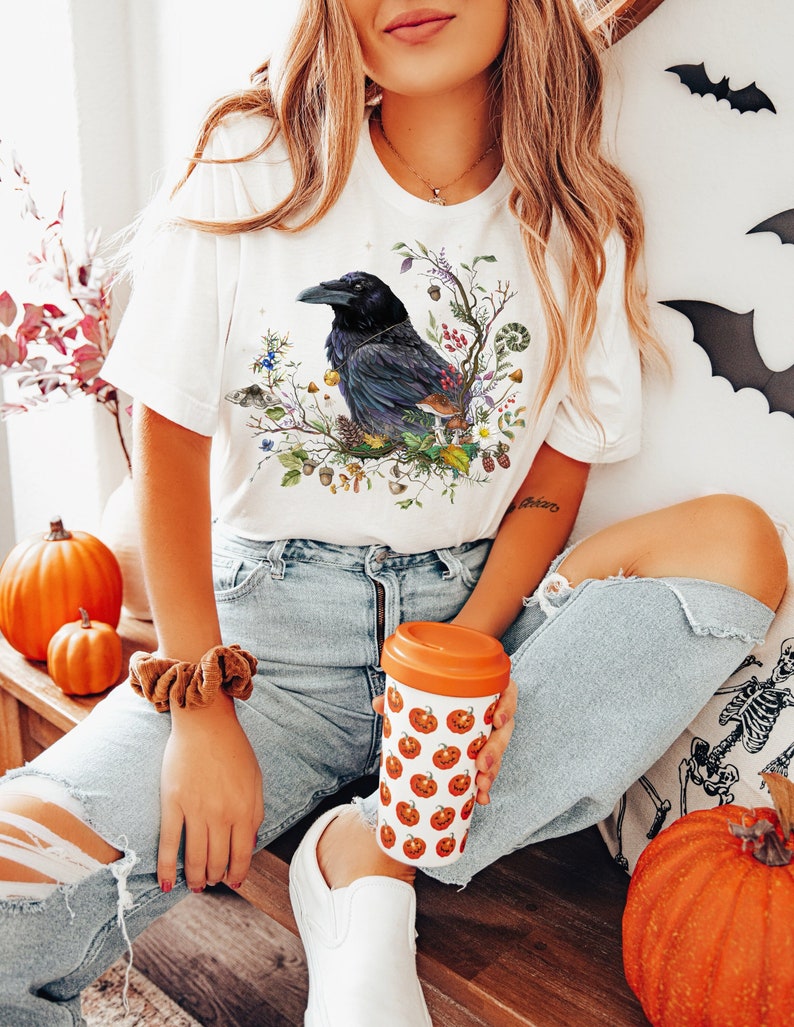 May include: A white t-shirt with a black raven illustration surrounded by autumnal foliage. The raven is perched on a branch with leaves, berries, and acorns. The t-shirt is worn by a person wearing blue jeans and a white shoe.