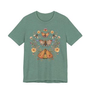 May include: A heathered sage green t-shirt featuring a central graphic print of three moths arranged vertically, surrounded by a circular arrangement of orange flowers, small berries, and green foliage in a soft, muted color palette.