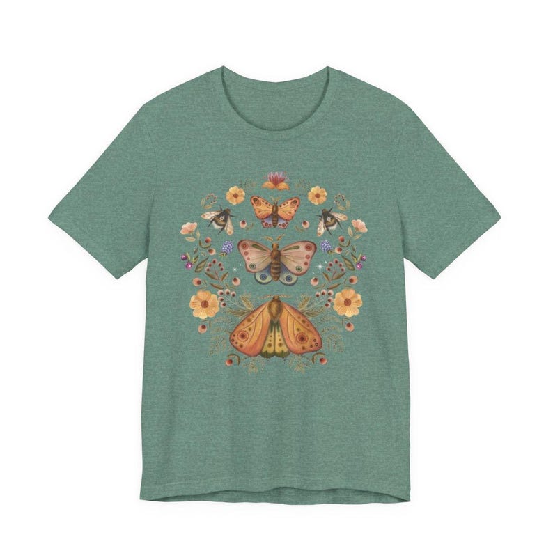 May include: A heathered sage green t-shirt featuring a central graphic print of three moths arranged vertically, surrounded by a circular arrangement of orange flowers, small berries, and green foliage in a soft, muted color palette.